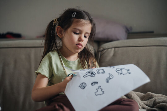 Young girl cutting paper with scissors holding sheet of simple doodle drawings on sofa in living room under soft natural light, focused expression, casual