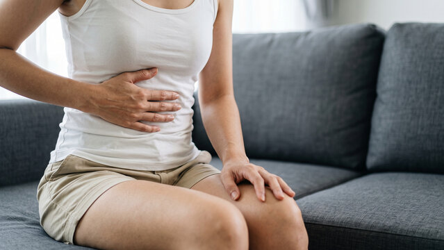 Woman Sitting on Sofa Holding Stomach with Abdominal Pain Showing Indigestion, Gastritis and Menstrual Cramps at Home