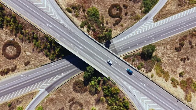 Cars travel on a highway interchange in a rural area. The road layout features multiple lanes and green patches nearby. The scene is captured from above in clear weather.