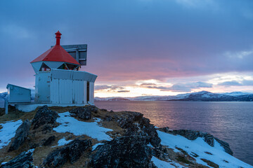 View of the Spildra lighthouse perched on a rocky, snow-dusted cliff overlooking the calm waters of a fjord during a colorful winter sunset in Kvaenangen Municipality, Troms, Norway. © Travel 'n' Lifestyle