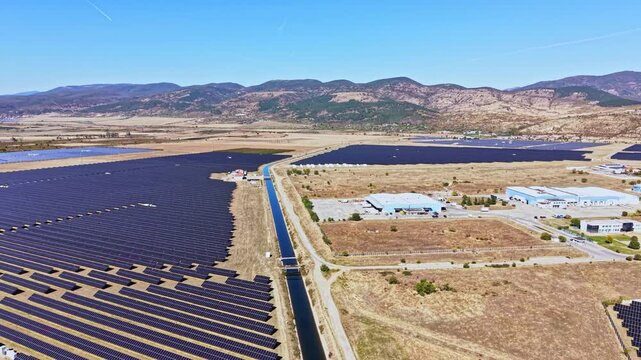 The scene shows a vast solar farm with rows of solar panels arranged in neat lines. The location is a valley surrounded by mountains on a clear day. There are roads and buildings nearby.