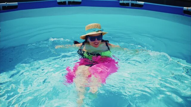Child splashes joyfully in bright blue pool. Wearing hat, sunglasses, and pink floaties. Smiling, playful, with arms raised mid-splash. Perfect for summer vacation, pool party, or fun activity themes