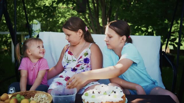 Three girls gather around a table outdoors. One girl smiles at the cake, others look on. The youngest reaches for fruit beside the dessert. Sunlight filters through the trees behind them