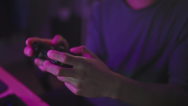 Man holding a controller with fingers on buttons at a desk with a keyboard and purple lighting in a studio gaming setup; concentration skill.