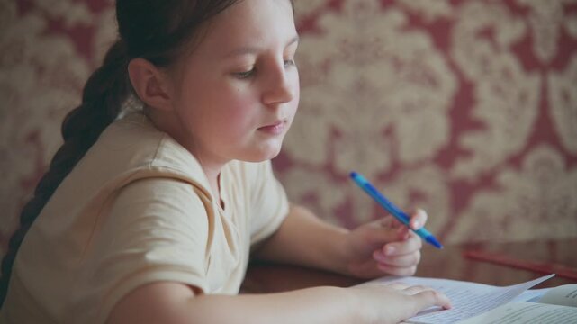 Young girl writes with blue pen at desk. She concentrates, eyes focused on paper. Background has soft, patterned wallpaper. Warm lighting enhances quiet, studious mood. Ideal for scenes of homework