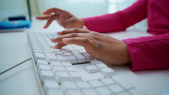 Hands type on keyboard, focused and steady. Pink sleeve suggests casual, productive work setting. Bright room shows calm, efficient computer use. Great for digital work, remote job