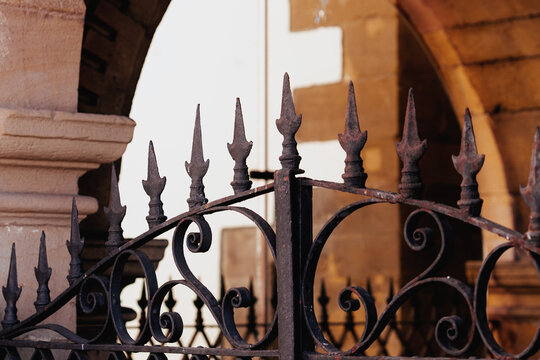 Decorative wrought iron gate spikes with intricate forged scrolls. Vintage rusted metal texture and ancient stone archway architecture in the blurred background