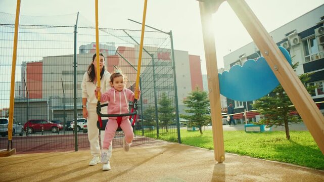 Soft sunlight bathes a wooden swing set. Mother smiles as she gently pushes a child. Child laughs, reaching for the swing's edge. Urban playground with green grass and buildings