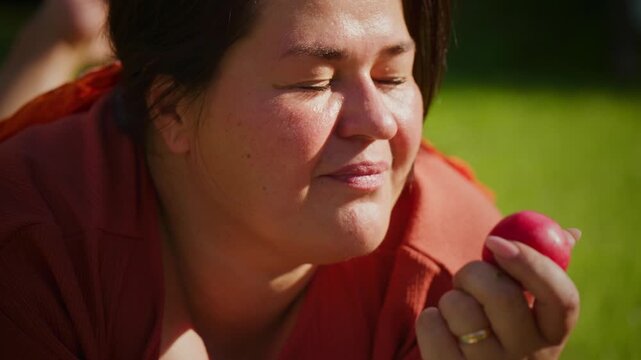 Woman bites red apple outdoors. Sunlight glows warmly on her face. She laughs softly, cheeks full. Green grass surrounds her gently. Ideal for healthy lifestyle or food-themed content