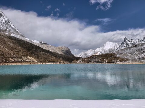 Crystal-clear turquoise alpine lake reflecting dramatic clouds and distant snow-covered mountain peaks in the high-altitude landscape of Daocheng Yading Nature Reserve, Sichuan, China.