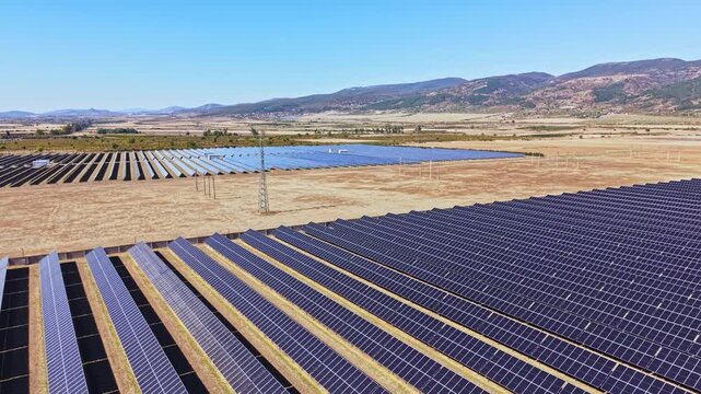 Large areas of solar panels are set in rows on open land. The bright sky shows no clouds. Mountains provide a backdrop to the fields.
