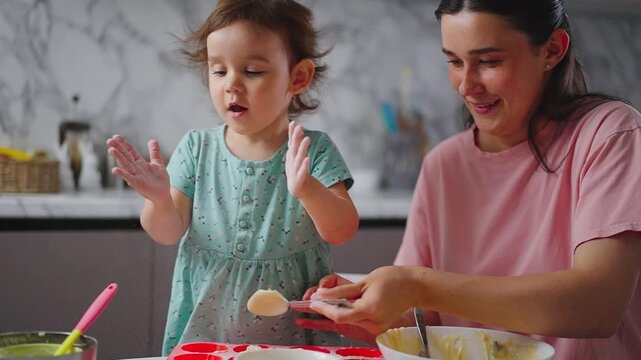 Toddler claps excitedly beside her mother. Mother smiles gently while stirring batter. They bake together in a bright kitchen. Perfect for family cooking, bonding, or parenting content