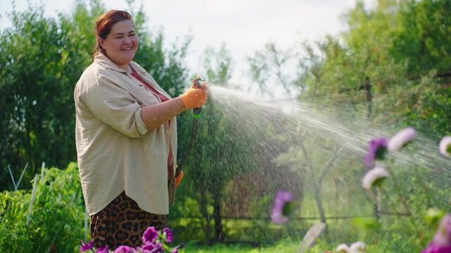 Woman waters garden with a cheerful smile. She wears gloves and a light jacket. Purple flowers sway in the gentle spray. Green trees frame the lush garden scene