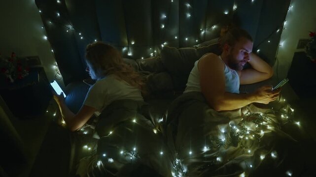 Soft lights drape the bed, casting gentle glow. Two people lie side by side, each absorbed in their screens. One person holds phone, eyes focused, fingers tapping. The other rests head