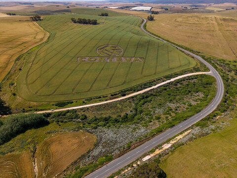 Aerial view of a large green field featuring a giant Corteva logo crop circle, surrounded by rolling hills and a winding asphalt road in the rural landscape of Caledon, Western Cape, South Africa.