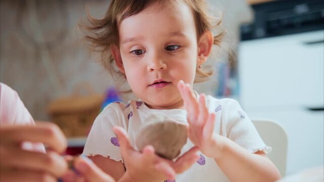 Toddler smiles while shaping clay with hands. Soft curls bounce as child focuses on play. Parent's hands guide gentle, nurturing interaction. Cozy kitchen setting enhances warm, creative mood