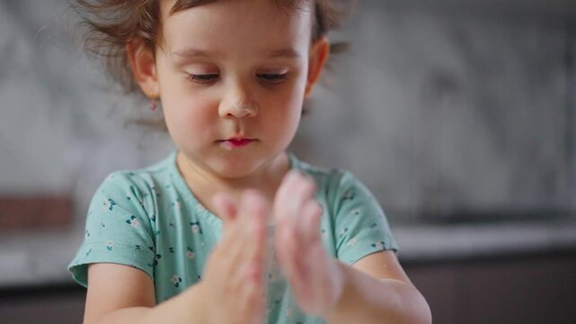 Little girl claps hands gently. She wears a green patterned shirt. Her focus is on the task at hand. Soft background suggests a kitchen setting. The scene captures innocent childlike joy