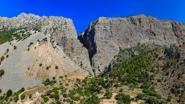Steep rocky cliffs rising above scrub-covered foothills and scree, tiny chapel visible near base of dramatic mountain fissure under clear blue sky