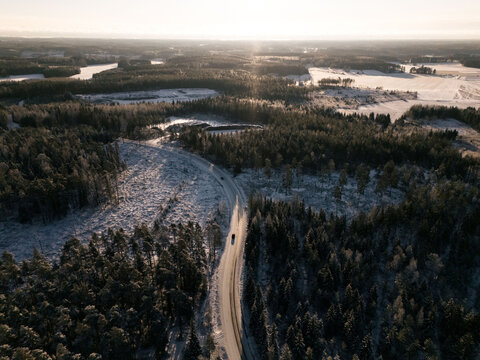 Aerial view of a winding road cutting through a snow-covered coniferous forest in the golden hour light in Uusimaa, Finland.
