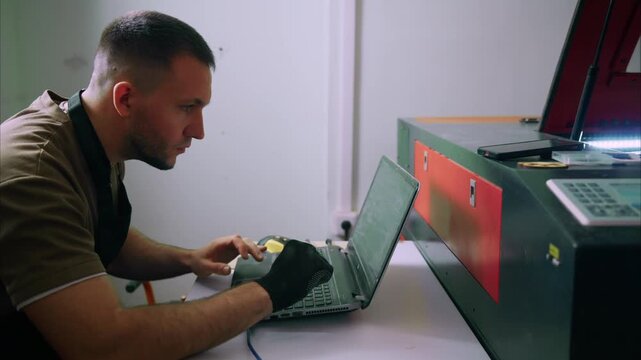 Man focused, typing on laptop beside industrial machine. He wears gloves, works intently, eyes fixed on screen. Equipment suggests manufacturing, fabrication, or engineering tasks