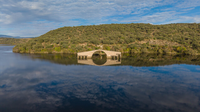 Aerial view of the ancient Roman bridge of Caparra partially submerged in the Gabriel y Galan reservoir, surrounded by hills and lush vegetation in Guijo de Granadilla, Extremadura, Spain.