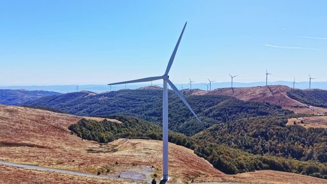 A wind turbine stands on a mountain with distant hills and a wind farm visible in the background. The sky is clear with no clouds.
