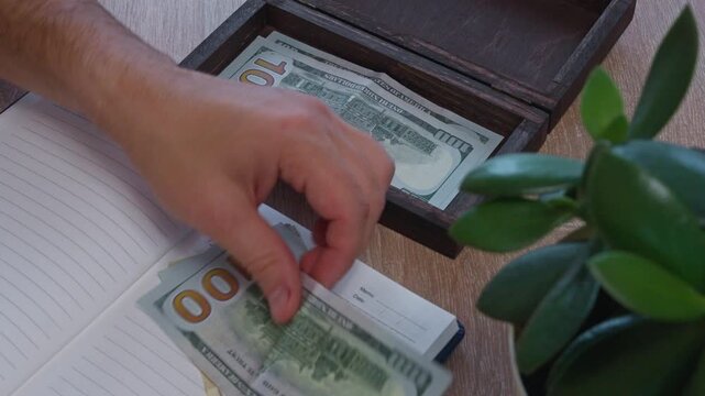 Male hands put dollar cash into wooden box establishing reserve fund, handling green banknotes near desktop plant. Methodical person manages domestic budget carefully, preparing safe nest egg.