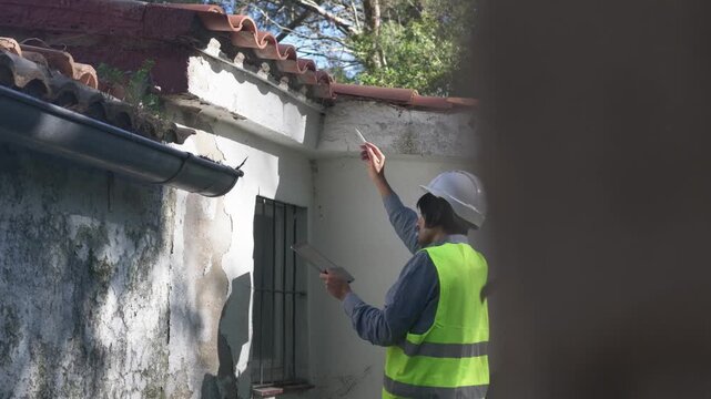 Building inspector wearing safety gear inspecting a damaged wall with a tablet. A construction engineer documenting cracks on an old building for structural analysis and renovation