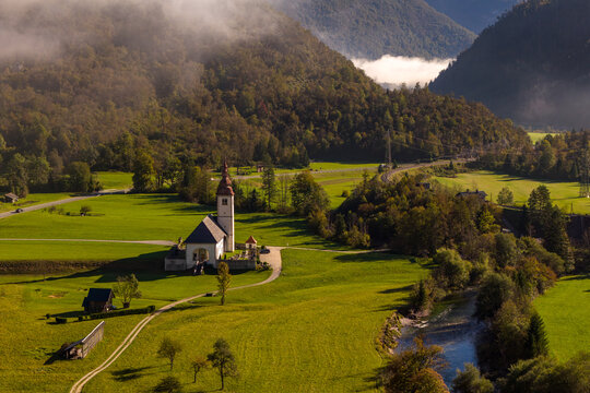 Aerial view of a historic church set in a lush green valley with a winding river, rolling hills, and morning mist in Bohinjska Bistrica, Radovljica, Slovenia.