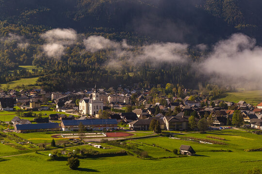 Aerial view of the town featuring a prominent church, surrounded by green fields and rolling hills under a misty morning sky in Bohinjska Bistrica, Radovljica, Slovenia.