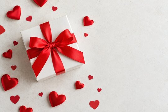 White gift box with red satin bow and small hearts on a light textured surface