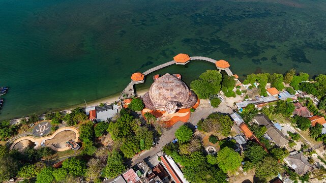 Aerial view of Kura-Kura Ocean Park, a turtle-shaped building, surrounded by lush trees, a wooden pier extending into the sea, and coastal structures in Jepara, Central Java, Indonesia.