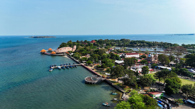 Aerial view of Kura-Kura Ocean Park with its turtle-shaped building, lush trees, coastal walkways, and boats moored in the clear blue sea in Jepara, Central Java, Indonesia.