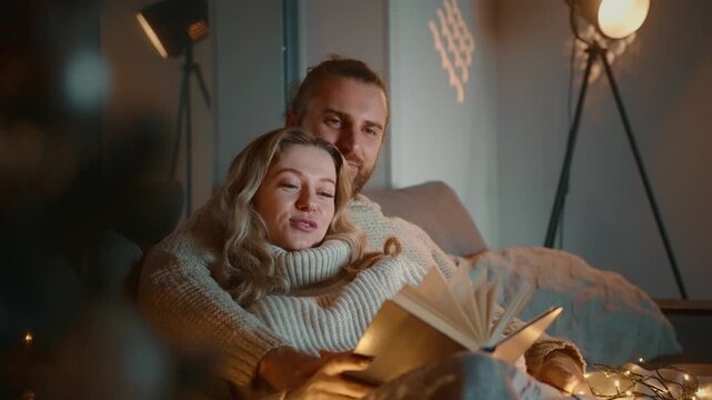 Cozy couple reads together in warm, soft light. They smile gently, sharing quiet, intimate moments. Bedroom features dim lighting and festive string lights
