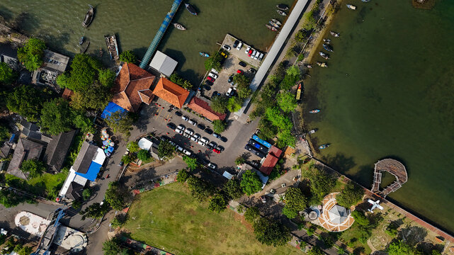 Aerial view of Jepara Kartini Ferry Terminal with a parking lot, traditional buildings with orange roofs, and boats docked along the pier in Jepara, Central Java, Indonesia.