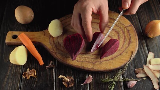 Hands slicing a red beet on a wooden cutting board surrounded by carrots, potatoes, onion, garlic, herbs, and other vegetables in a top view kitchen scene