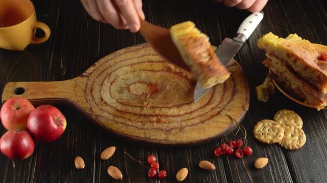 Hands slice a piece of layered cake on a rustic wooden serving board with a knife and spatula, surrounded by apples, crackers, almonds, berries, and tea