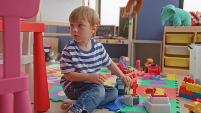 Boy sits crosslegged playing with toy cars and stacking blocks on a colorful foam mat amid storage shelves and plush toys in building; curiosity.