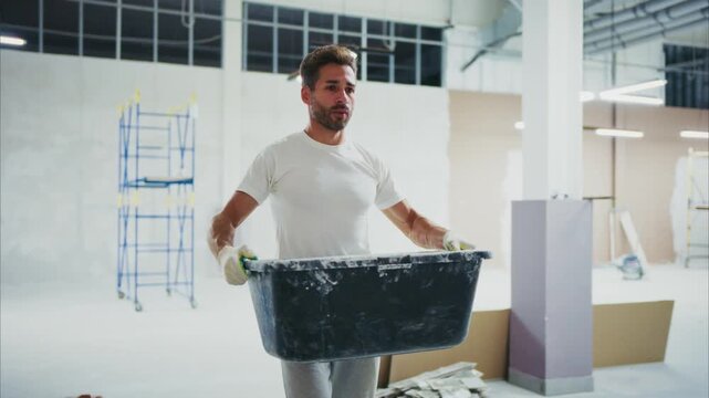 Man carries paint container in bright, spacious studio. He bends to dip brush into mixing tub, focused. Workshop walls show exposed drywall, tools nearby. Ideal for DIY, home renovation