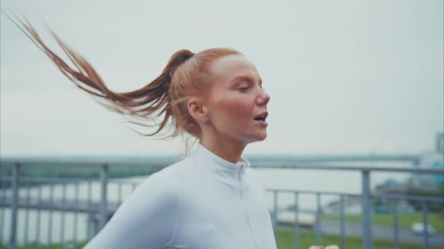 Woman runs outdoors, wind whipping her hair. She moves steadily, eyes closed, focused on pace. Light gray sky frames urban waterway behind her. Feeling calm, she breathes deeply, enjoying motion