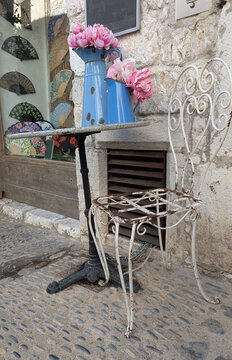 Vintage wrought iron chair and pink flowers in blue enamel pitchers outside a shop in Tourrettes-sur-Loup, France. 