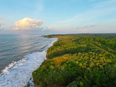Aerial view of Pantai Pasir Besi featuring lush green coastal forests and rugged cliffs meeting the ocean waves under a clear sky, Cibitung, West Java, Indonesia.