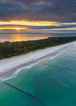 Aerial view of Hel Peninsula at sunset with sandy beach, turquoise sea, dark forest, and wooden groynes in Chalupy, Pomeranian Voivodeship, Poland.
