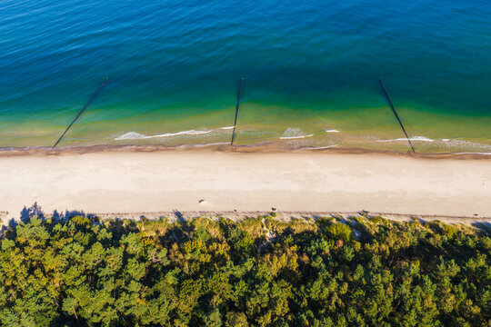 Aerial view of Hel Peninsula featuring a sandy beach, turquoise sea with wooden groynes, and a dense pine forest in Chalupy, Pomeranian Voivodeship, Poland.