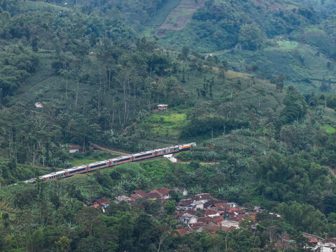 Aerial view of a passenger train traveling through a lush, hilly landscape with dense vegetation and a small village in Kadungora, West Java, Indonesia.