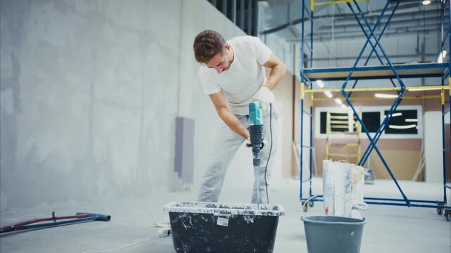 Young man mixes concrete with electric tool. He concentrates, focused on mixing task. Bright space with scaffolding and buckets nearby. Ideal for construction, renovation, or DIY projects