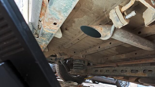 Exhaust pipe of a running car emits smoke and vibrates under the vehicle chassis during daytime, close-up undercarriage view.