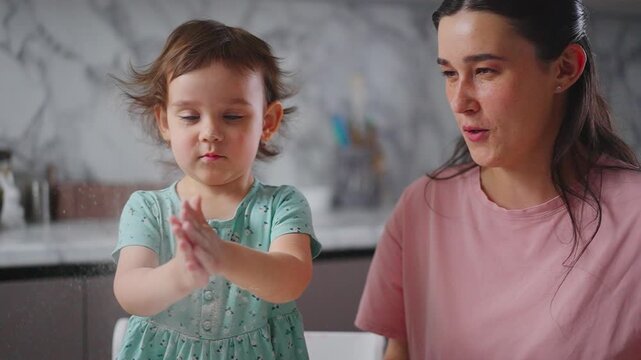 Mother and child share joyful kitchen moments. Child claps hands, smiling brightly. Mother watches with warm, amused expression. Ideal for family, parenting, or health care content