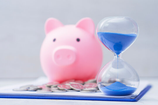 Hourglass with blue sand standing in front of pink piggy bank and stack of coins on clipboard. Time is money, financial planning, investment growth, retirement savings and deadline concept.
