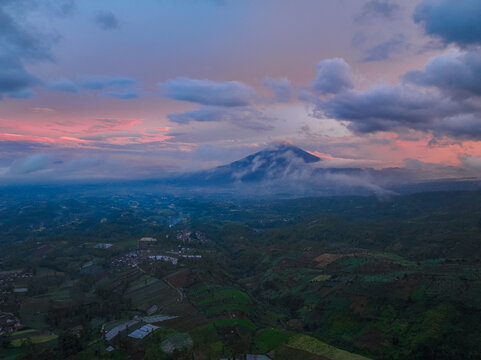 Aerial view of Darajat Pass at sunset, featuring a mountain peak partially shrouded in clouds above lush agricultural fields and rolling hills in Kecamatan Pasirwangi, Jawa Barat, Indonesia.